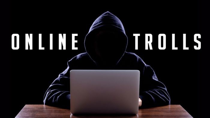 Dark and eerie photography of a person in a hooded top with no visible face, sitting at a desk in front of a laptop computer. Behind them in large white letters spells ONLINE TROLLS