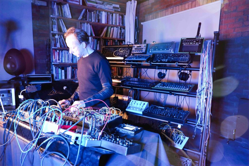 Colourful image of a music studio, with musical equipment, lights and a a table set up with music gear on it. A man stands in front performing at a large table of electronic equipment