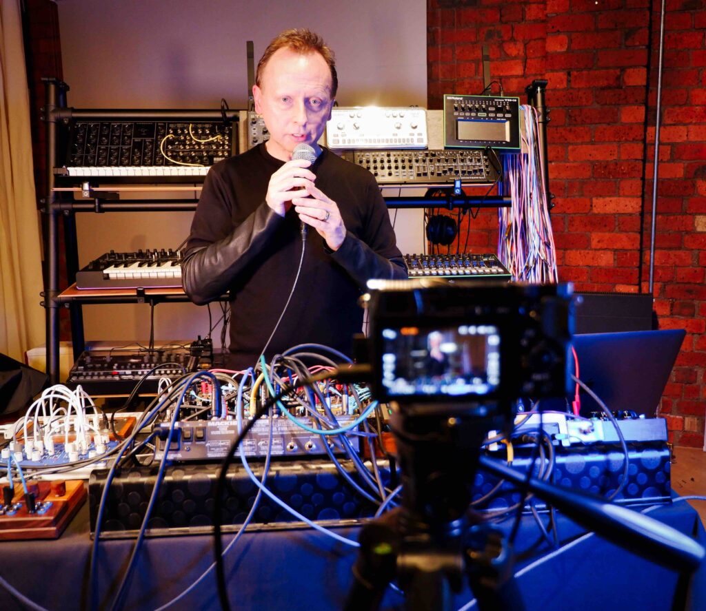 A man stands holding a microphone looking at the camera, surrounded by musical equipment in a music studio. He is dressed all in black from the waist up