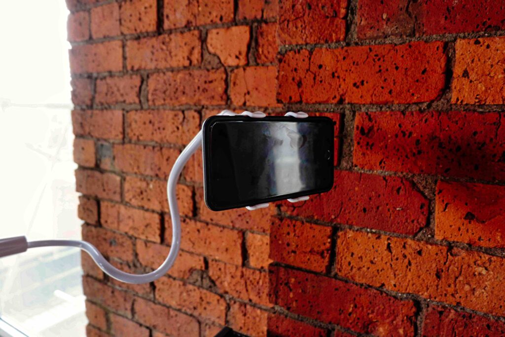 An Apple iPhone is held up in the air by a white stand, pictured against a red brick wall