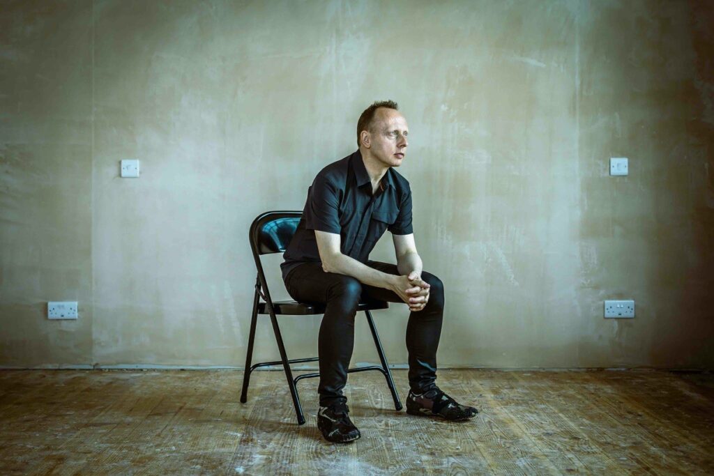 A male figure, Robin Rimbaud, sits on a metal black chair in an industrial looking room, otherwise empty. He wears a smart black shirt unbuttoned at the collar, and tight black shiny trousers, and sneakers. He stares in the distance. Four plug sockets are visible on the wall behind him.