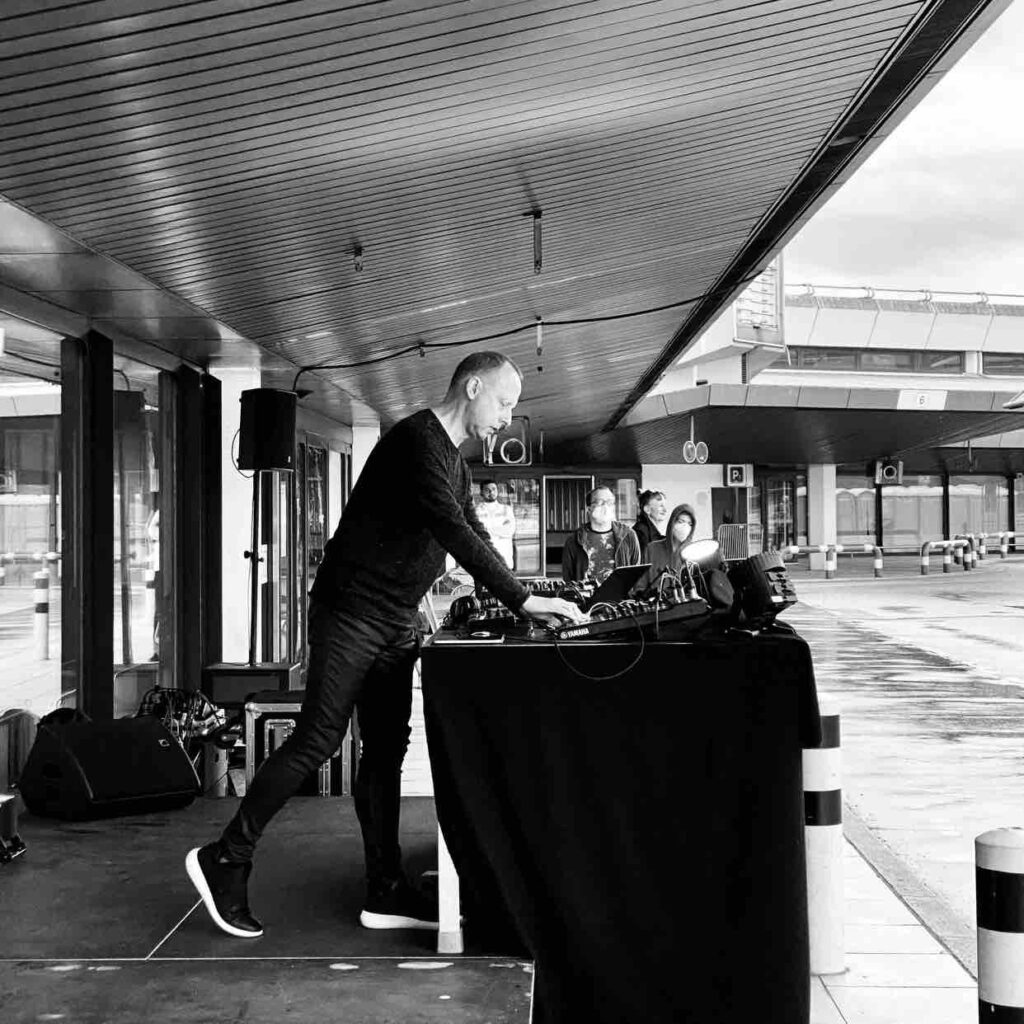 Black and white image of Robin Rimbaud, an older white man, in tight black coated jeans, shoes with a white layer at the bottom, black jumper. He's leaning over a large table with a bank of electronic equipment performing outdoors at Tegel Airport in Berlin