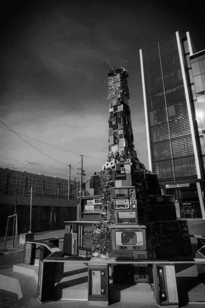 Black and white image of a large bank of old tv sets, radios, and electronics all constructed into a giant tower in a city