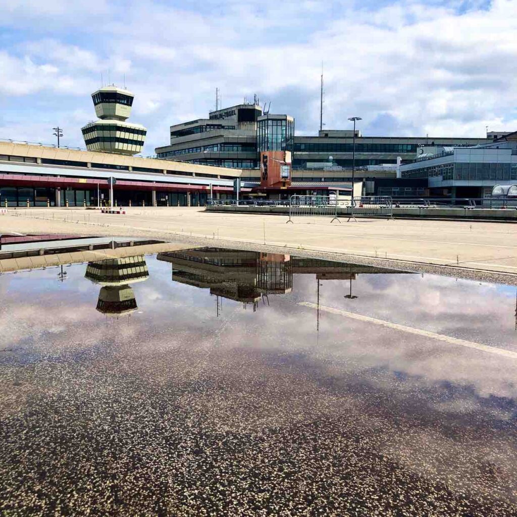 Colourful image of Tegel Airport in Berlin, devoid of people and travellers. You see a reflection of the building in a large rain puddle on the ground