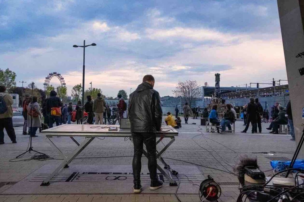 Man seen from behind standing at a table outdoors performing electronic music. A large crowd of people face away from him looking into the distance. He wears a shiny black jacket and skintight black shiny trousers