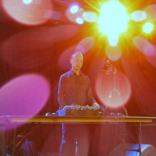 Photograph of a man in a very smart black shirt, standing on a stage in front of an electronic synthesiser, looking out into the audience. The stage lights are bright and glaring all around him