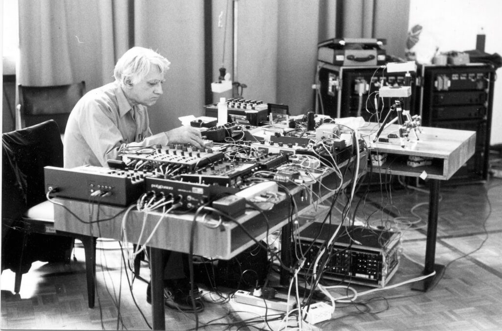 Black and white photograph of an older white man, with white hand wearing a white shirt seated in front of a large table. The table top is completely filled with strange anonymous boxes with wires and cables everywhere
