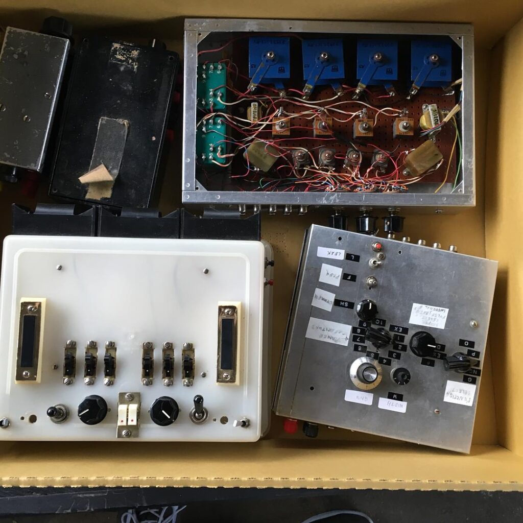 Four metal boxes sit inside a cardboard box. Each displays switches and buttons and one has homed made labels stuck to the surface of it