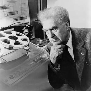 Edgard-Varese Black and white photography of elders man, with grey hair, in a smart black suit, leaning on his hand in though in a music studio. Behind him stands a old reel-to-reel tape player