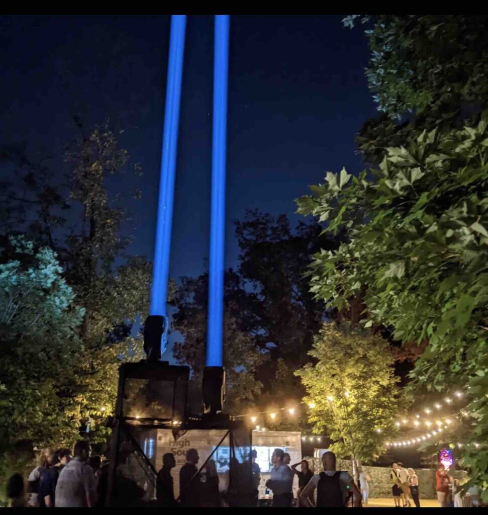Two strong beams of blue light shoot in the sky from the ground in an outdoor scene. Trees and foliage can be seen all around as a crowd of people stand around the bottom of the projector