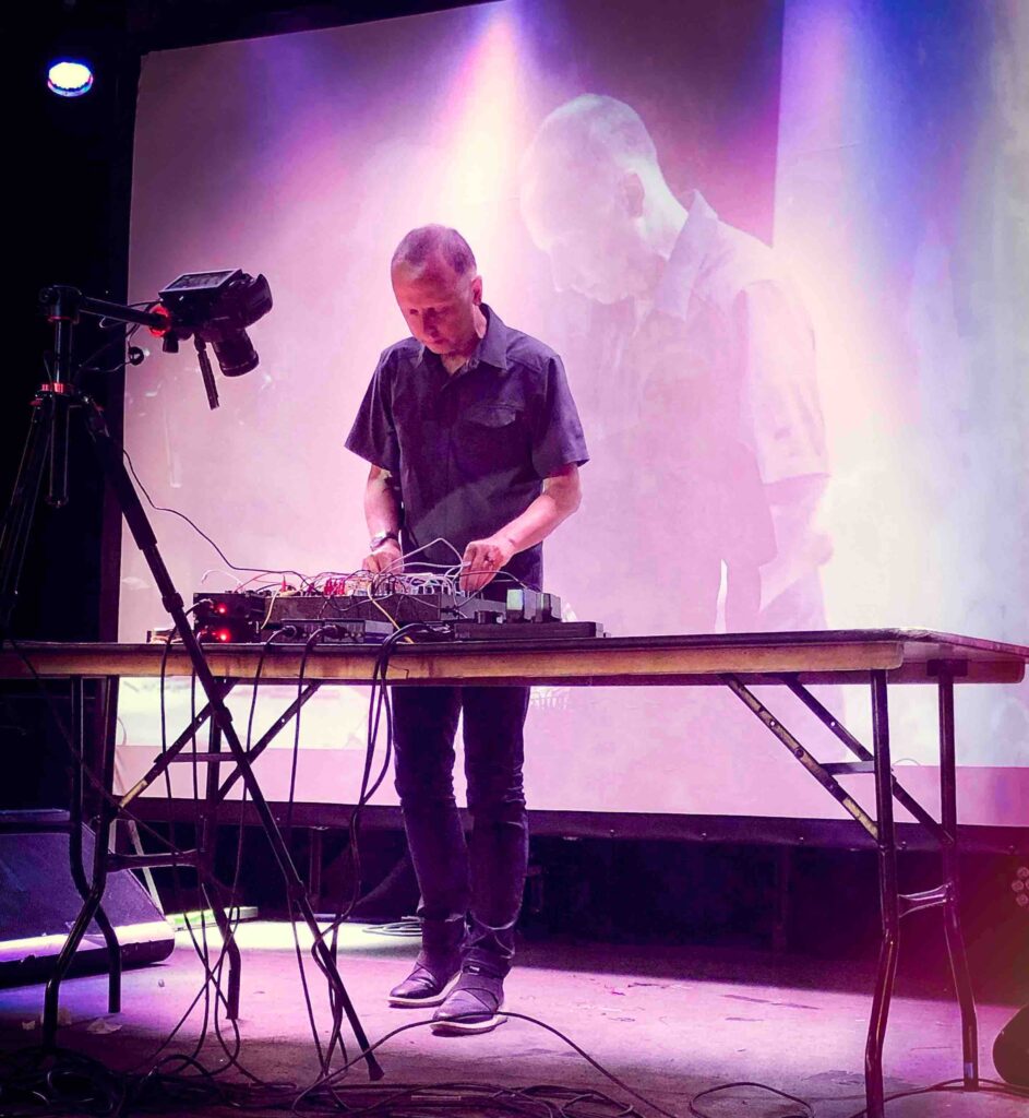 A man in a smart white shirt stands at a table, staring down at a modest bank of equipment, on stage. His image is also projected behind onto a large screen and another camera can be seen filming the equipment