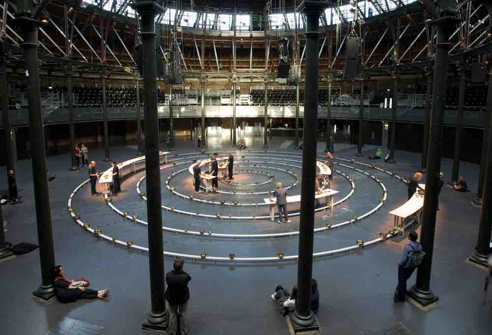 Inside the Roundhouse building in Camden London, you can see a series of concentric circles with people standing at wooden tables in different parts, with spectators standing around the outside listening to the music