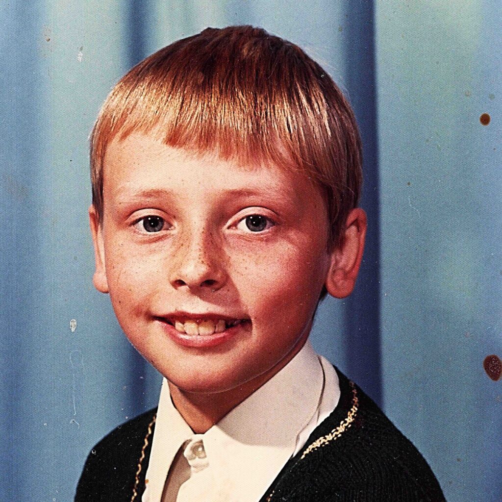 A young boy with blonde hair, aged 11 years old, smiles at the camera, wearing a smart shirt and jumper, with a blue background behind him