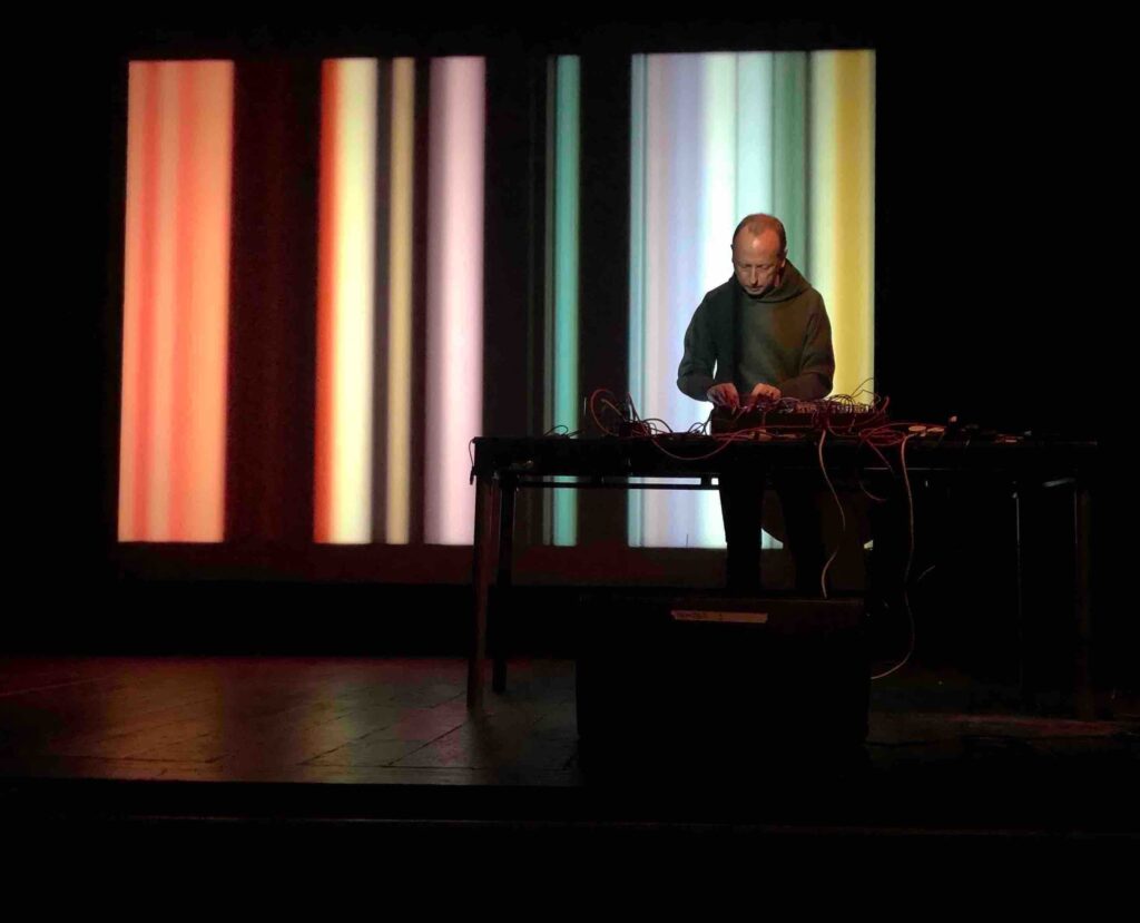 A white man stands at a table on a stage leaning leaning over a bank of equipment, wearing a hoodie. Behind him a series of bold colourful stripes are projected onto a screen