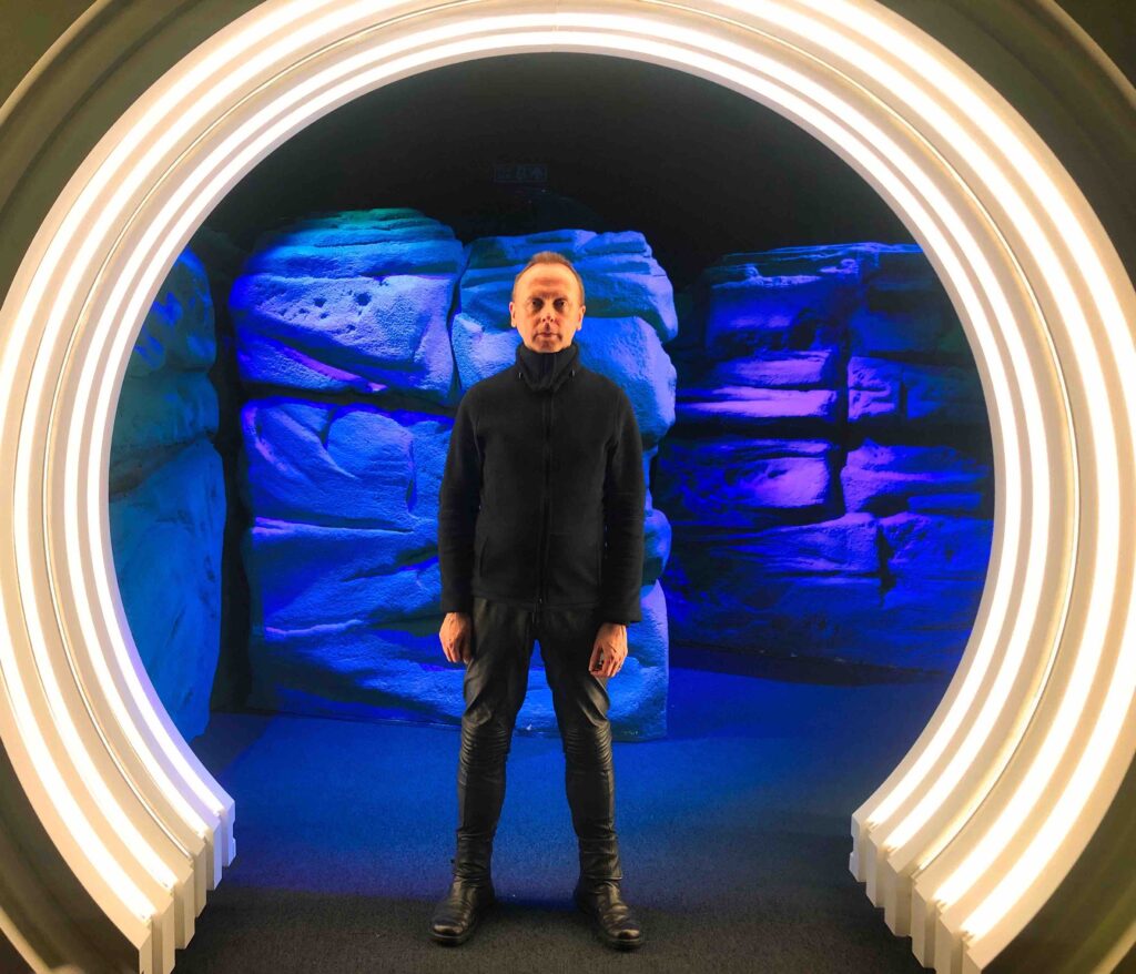A white man dressed all in black, in leather trousers, stands in the middle of a large white circle of light, with blue rocks lit behind it