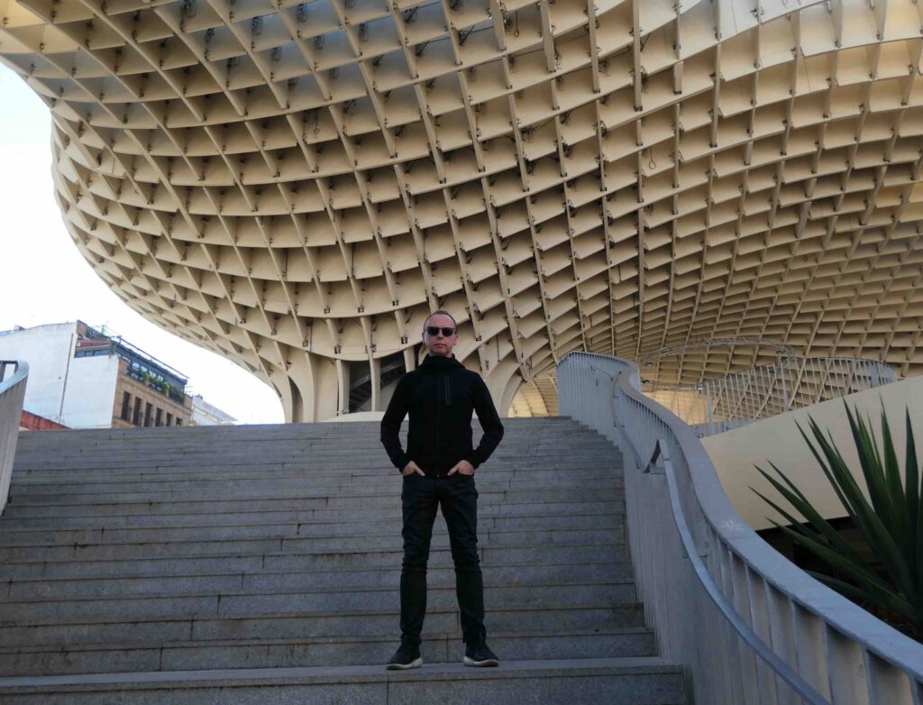 man with short hair dressed in slim black clothing stands on a concrete staircase, beneath a huge wooden structure, looking very alien like, modernist architecture prevailing