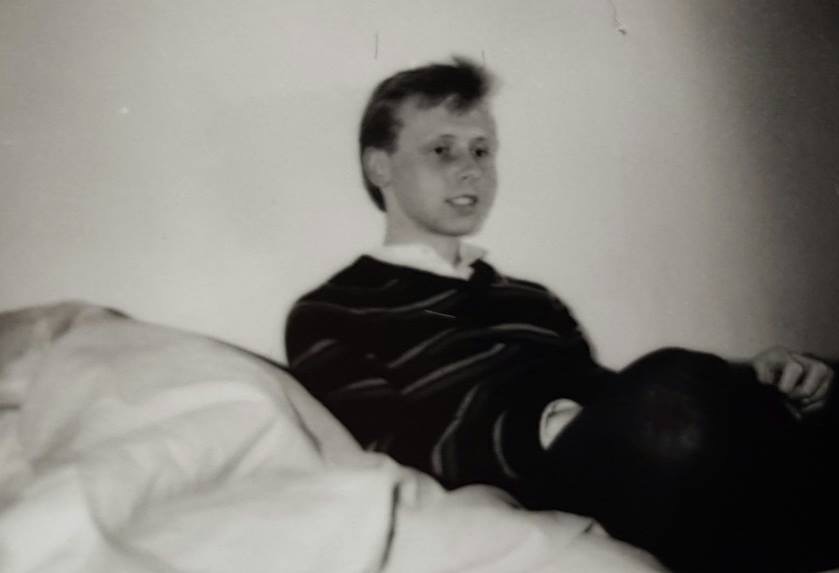 Black and white photograph of white boy, teenager, laying on a bed, wearing a striped jumper and white shirt
