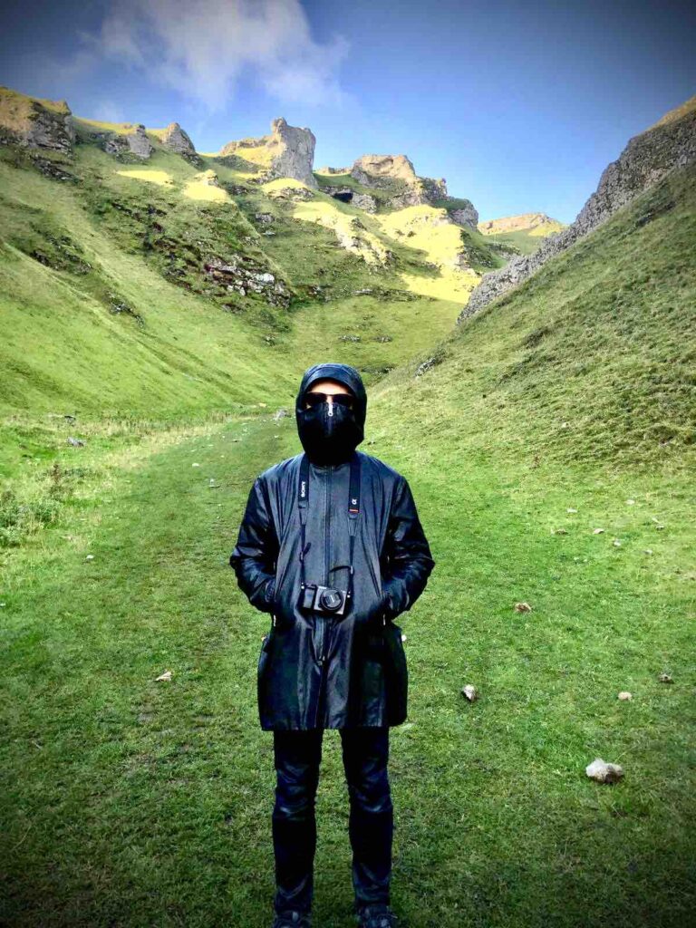 The Peak District in the UK, with a man standing there completely obscured by a long black coat, wearing sunglasses, standing in between two huge hills