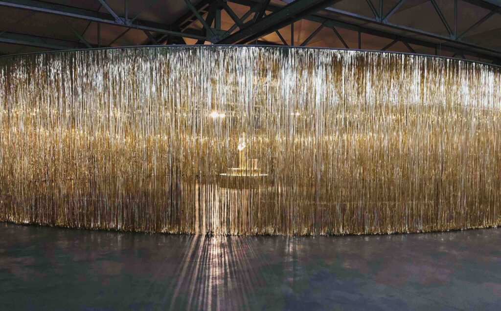A huge golden curtain hangs up in an art space, with light shining through it
