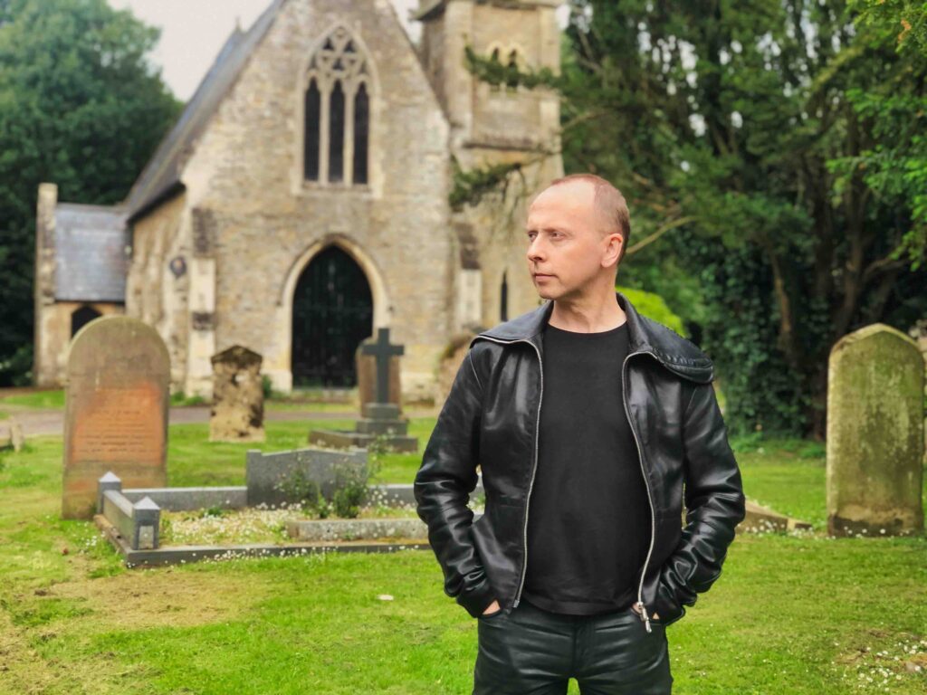A handsome white man with short blond hair stands in a graveyard looking across to his right, wearing a leather jacket and tight leather trousers, with a moody atmosphere