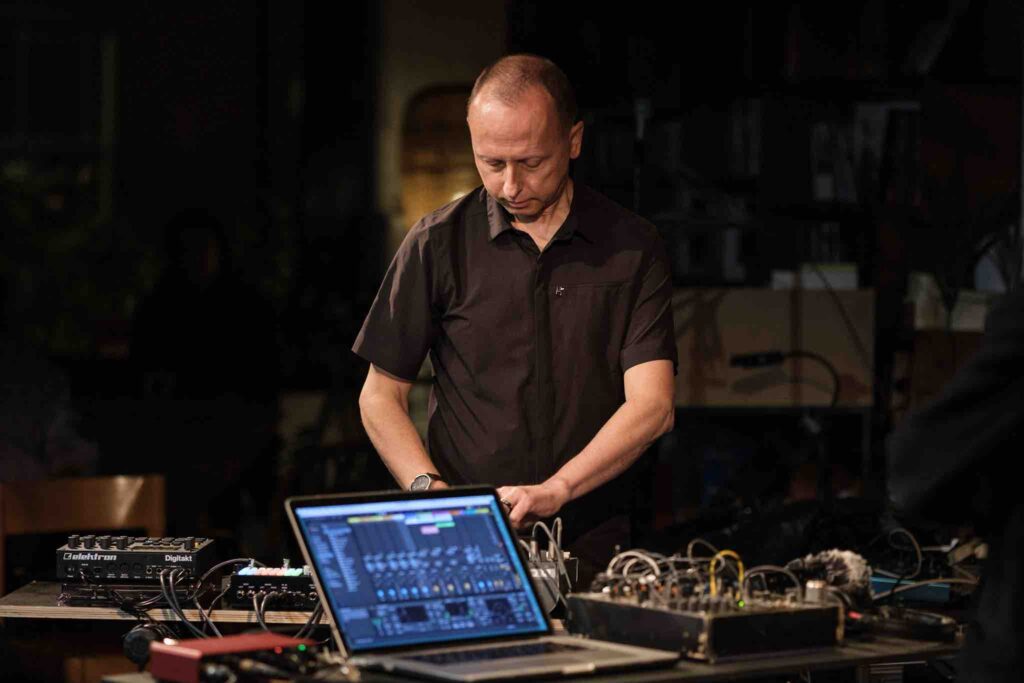 A white main a smart black shirt stands at a table, with musical equipment, a laptop in a performance space