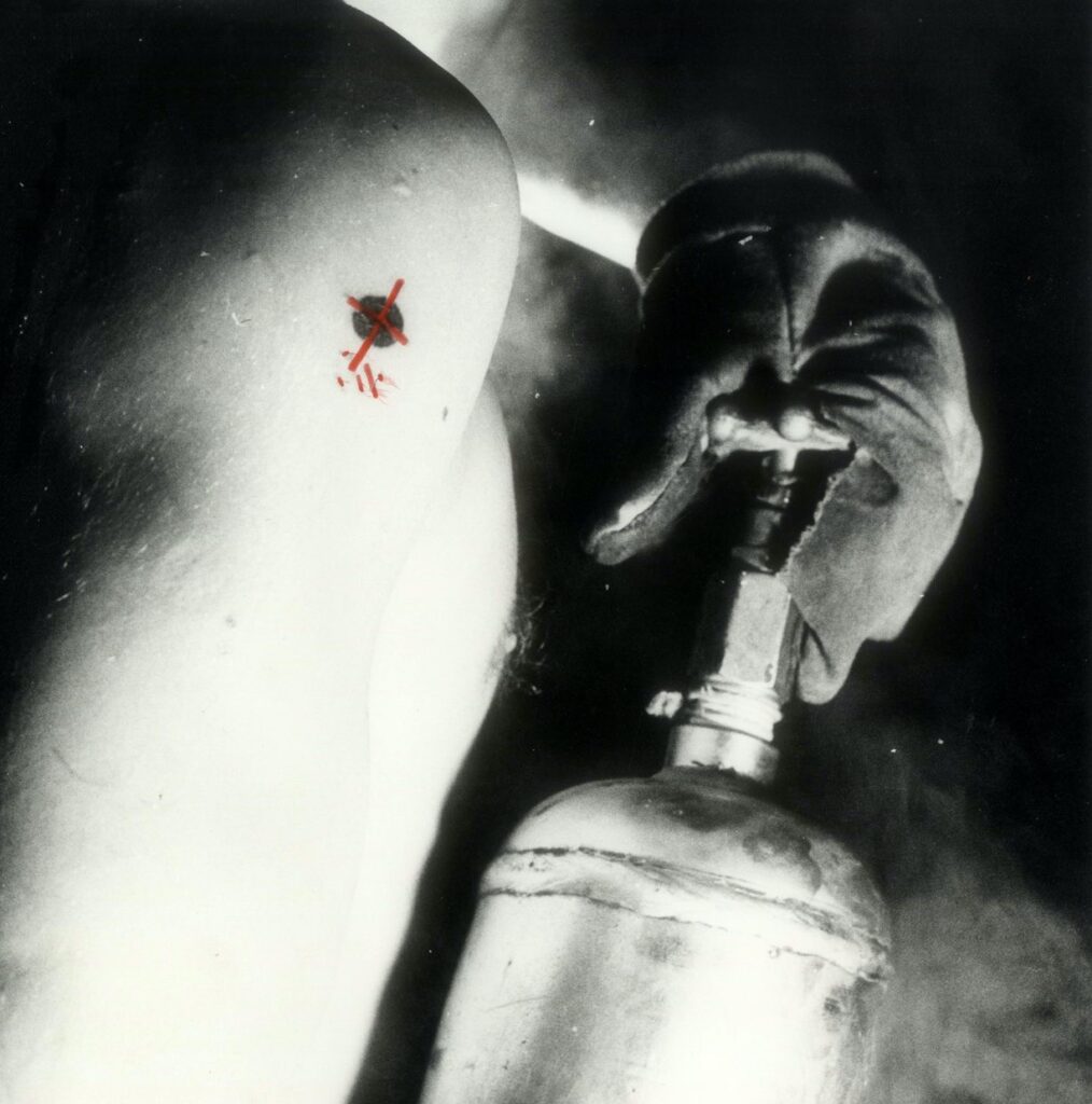 Black and white image of a man's naked shoulder. You can see a gloved hand pushing down on a valve over a fire extinguisher. The shoulder displays a tattoo of a black circle with a red cross over it