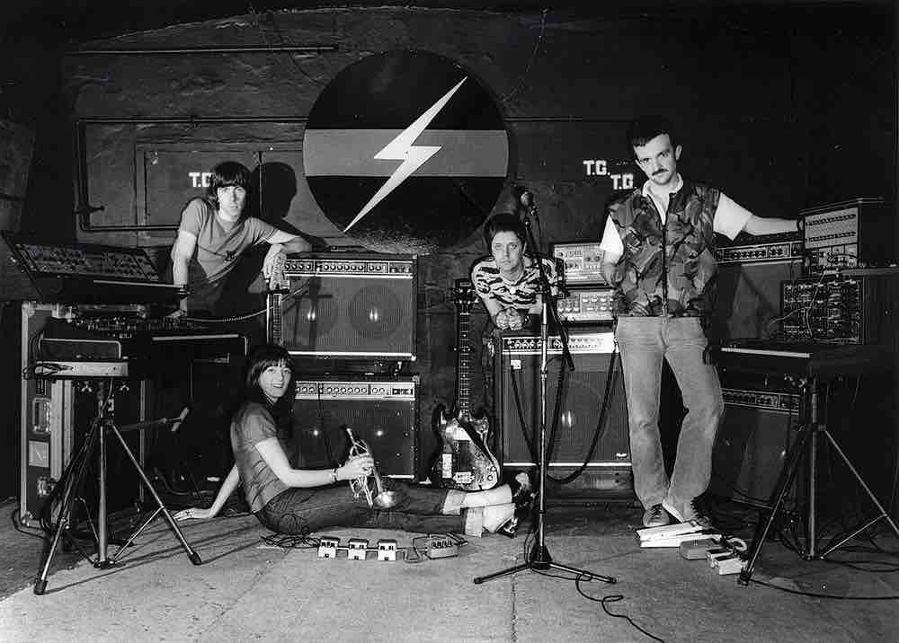 Black and white photo of British industrial music band Throbbing Gristle with four people, 3 men and 1 woman, standing in a studio, around a bank of guitar amps and instruments