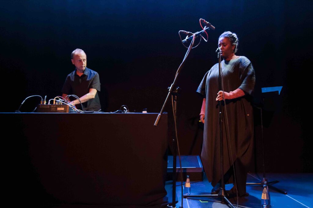 A white man stands at a table on the left on a stage, with a bank of electronic equipment in front of him. He stares at a larger black woman to his left who is singing into a microphone, dressed in a long black dress