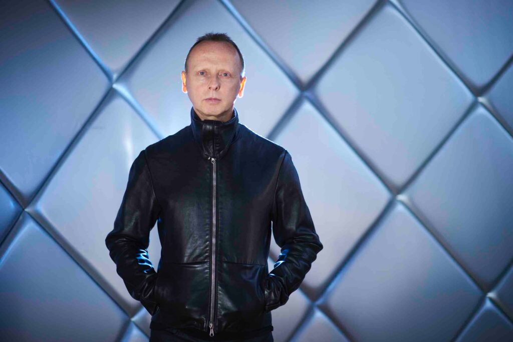 Image of man in his 50s, wearing a skinny leather jacket zipped up to his neck. Short crossed hair and staring directly at the camera, cool, no smile. Hands in pockets of jacket. Behind him is a shiny padded wall made of of angled squares, with light coming from behind him