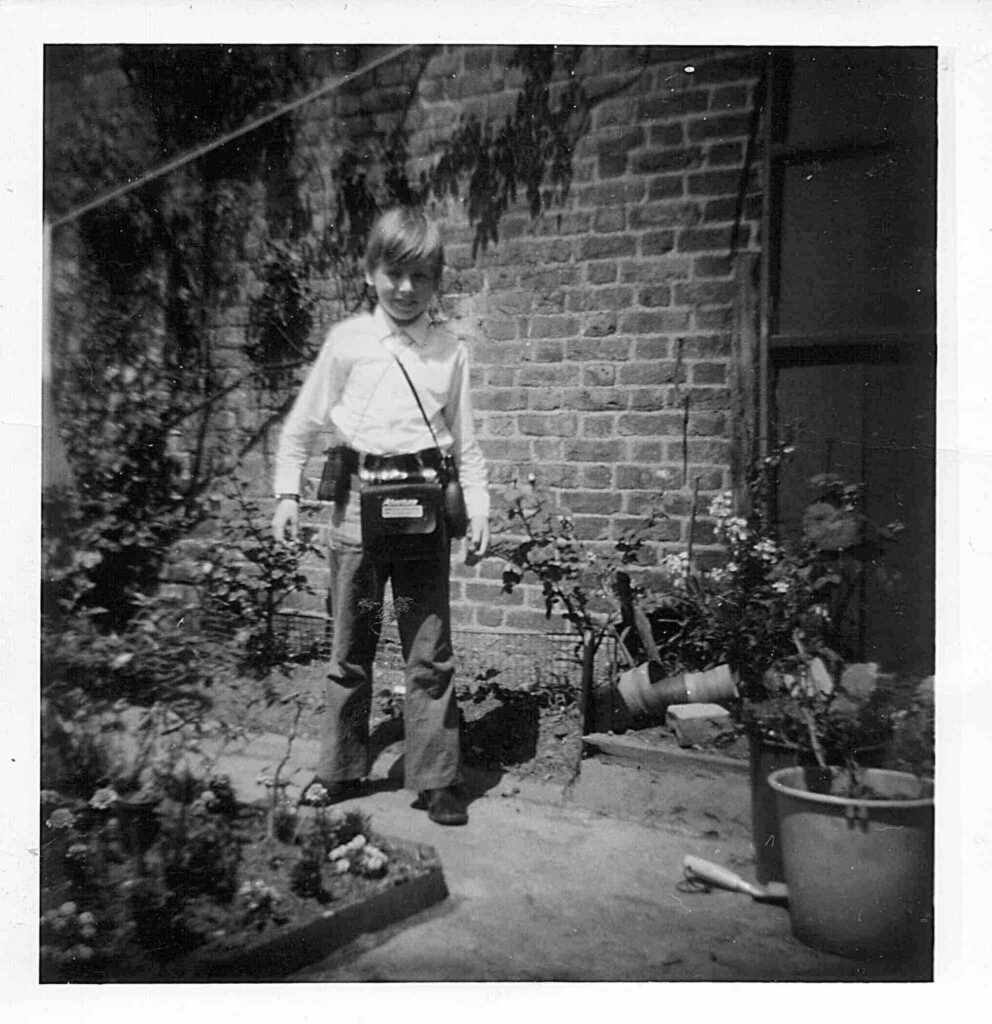 Black and white photograph from 1970 of a young boy aged around 8 standing in the garden, wearing a white shirt and slightly flared jeans. He's got blonde hair over his forehead and wears binoculars and other items around his neck
