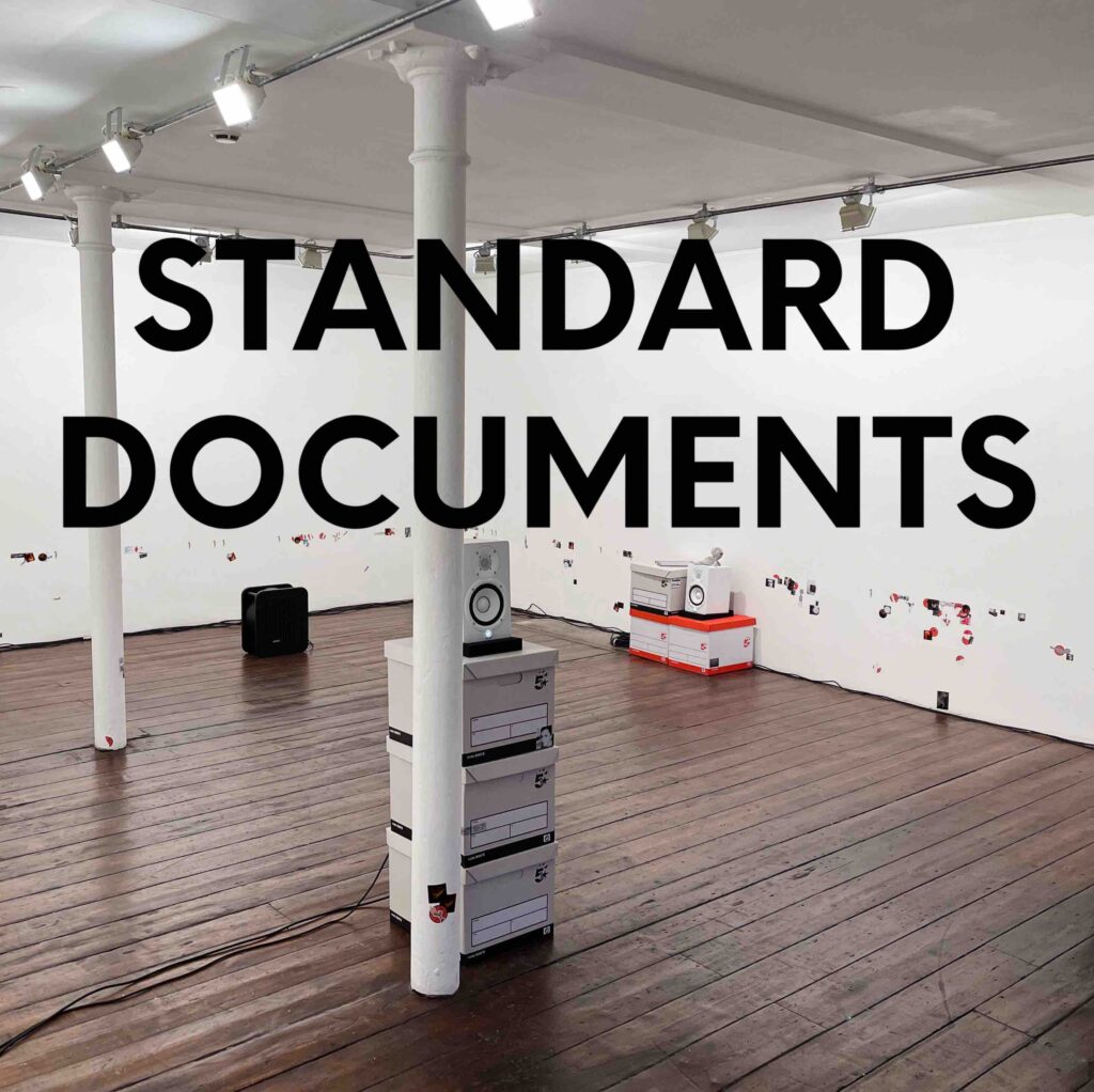 A gallery space, with wooden floor and white walls. Stacks of office papers sit on the floor with speakers on top of them. A large bold typecast across the image says 'standard documents'