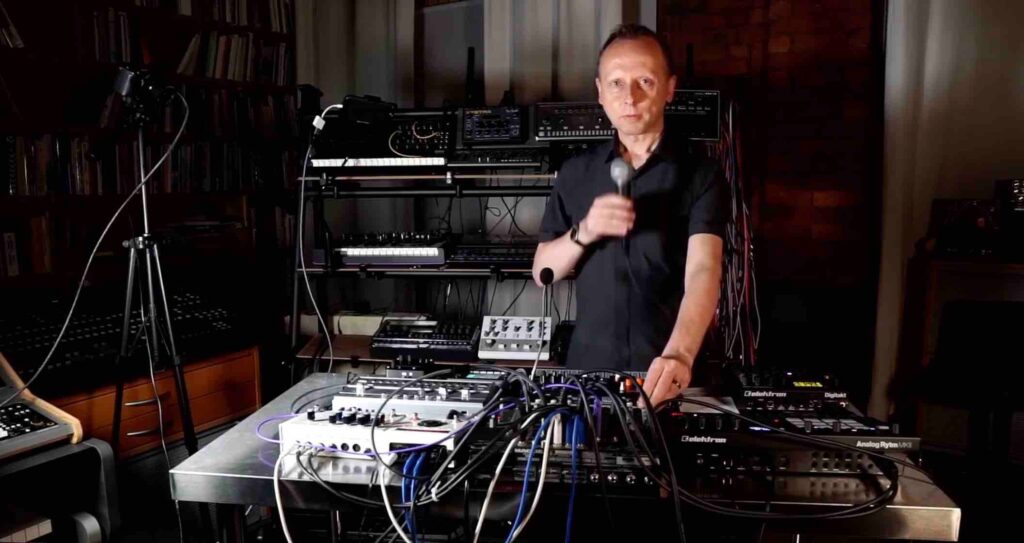 A very handsome man with short hair stands in a music studio holding a microphone. He wears a black shirt and stands behind a table filled with electronic music equipment as he stares at the camera.