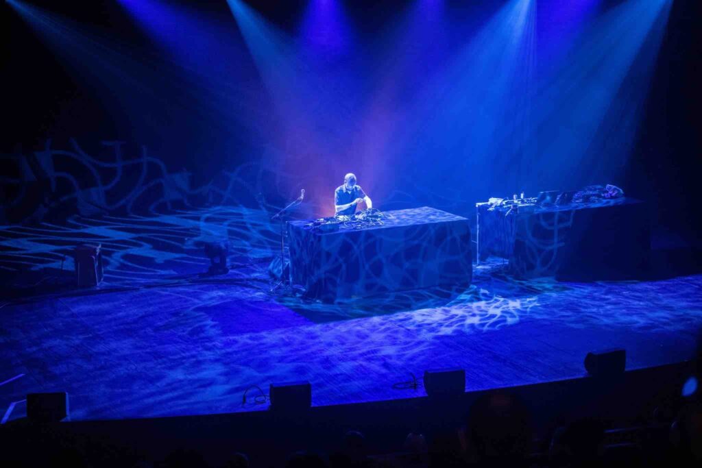 Dramatic image of a man on stage covered in blue light. Photo taken from audience so you can see his standing at a table in front of a bank of electronic music equipment.