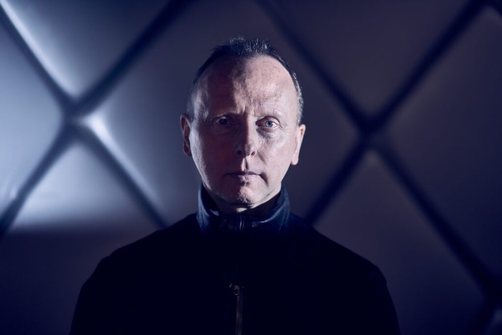 Image of man, aged around 50, serious, staring directly at the camera. Wearing a high neck leather top he has short hair and an intense stare. Behind is an abstract padded wall looking like a cross
