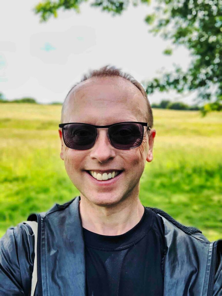 Colourful image of a handsome white man wearing sunglasses, with cropped blonde hair. A wide smile, leather coat, with the countryside behind him