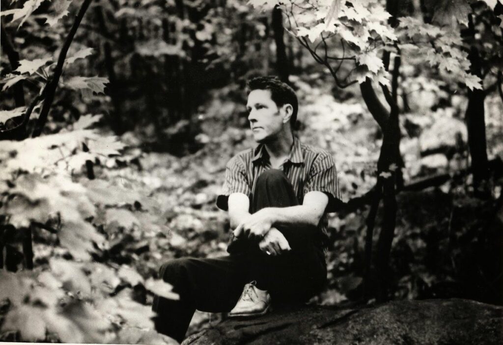 Black and white photograph of a man, the American composer John Cage, sitting in a forest, casually dressed, staring thoughtfully into the distance