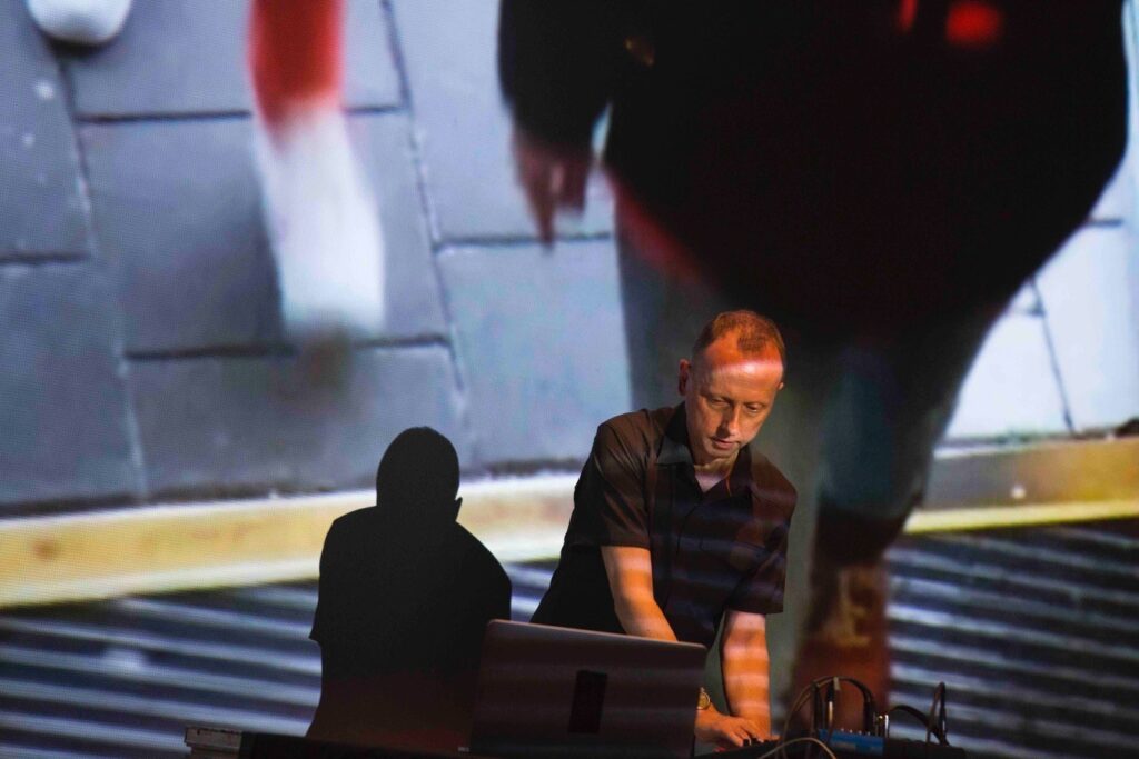 Photograph of a short haired man, Robin Rimbaud, standing at a table, wearing a black shirt, leaning over a computer, in a live musical performance, with a huge projection screen behind him, in a very dramatic pose