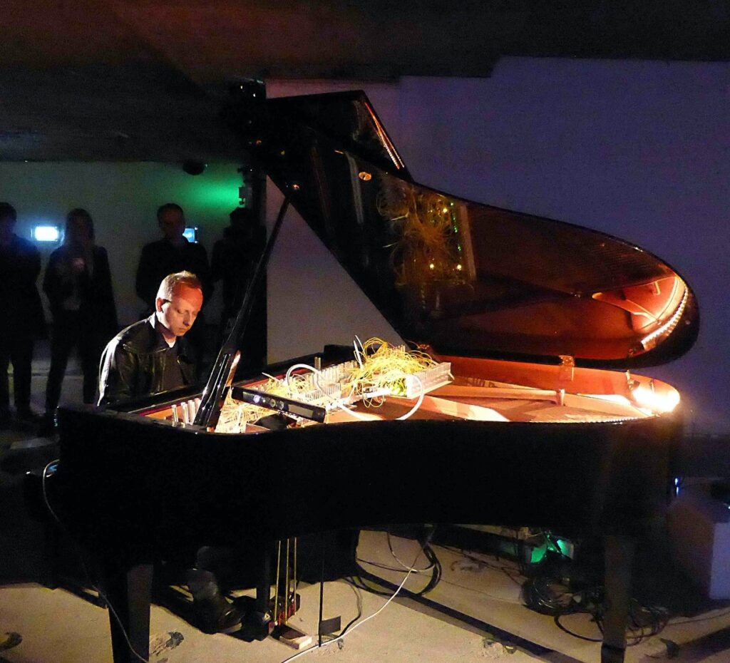 Photograph of a man in leather jacket sitting playing the piano. The piano has all kinds of wires and cables inside it, There are people standing in the room behind the pianist, in the dark
