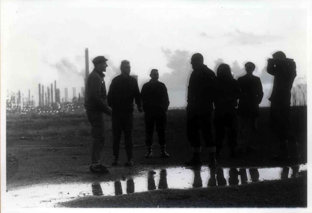 Black and white photo of seven figures, all in shadow standing outside in a gloomy wet industrial area.