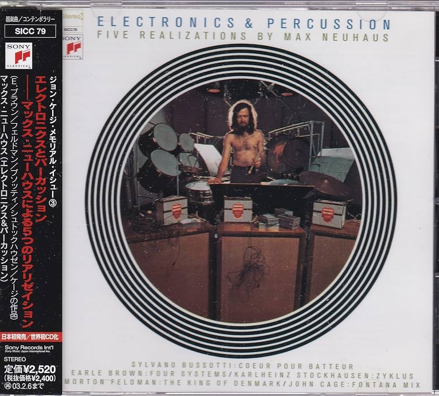 A vinyl LP record sleeved with a topless man, in brown trousers, standing in the centre of a giant circular image, surrounded by percussion equipment, holding a drum stick. Huge amplifiers stand at the front of the photo