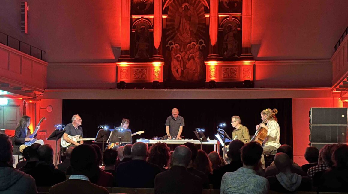 Photo of group of musicians on a theatre stage, with red lights illuminating them. Musicians playing guitars, cello, and electronics