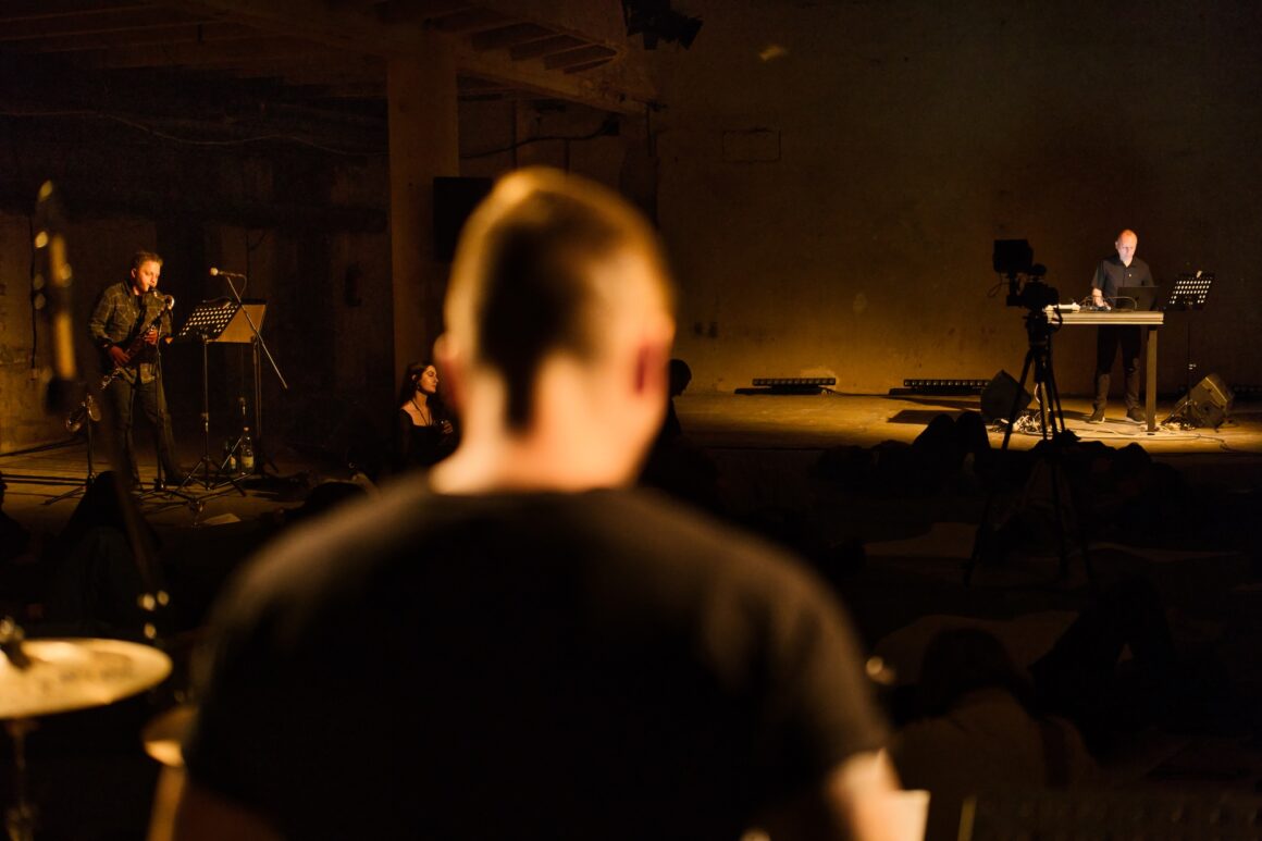 A blurred figure seen from behind features in the image of a live performance, with a man playing clarinet on his left and in the distance a man standing behind a table