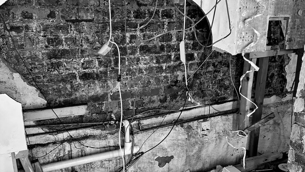 Black and white photograph of a brick wall inside a house, with hanging cables and pipes, with a real sense of destruction about it