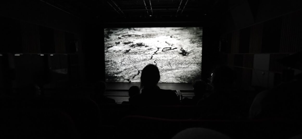 Atmospheric photo in black and white of a couple of people standing in front of a film projection in a very dark space.