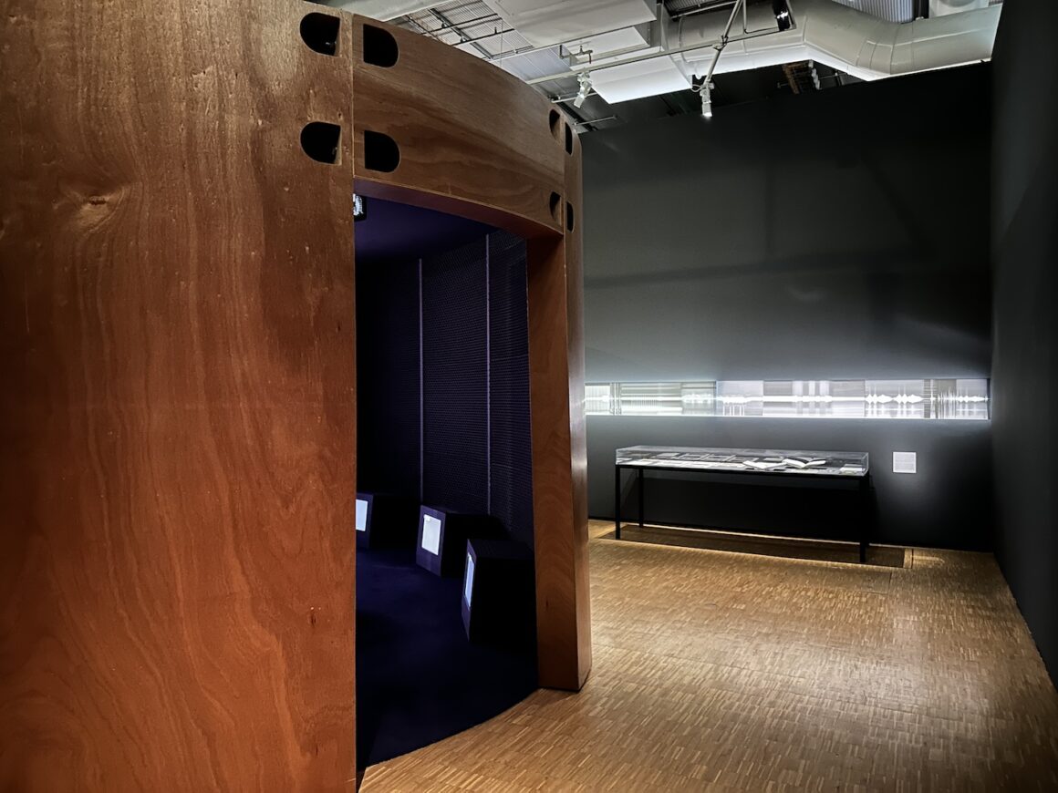 A large wooden structure with an entrance is on the left of the picture, with small tv screens seen on the inside of the structure. On the right is a large glass museum case displaying items which can't be made out. Light glows around the wall and wooden floor