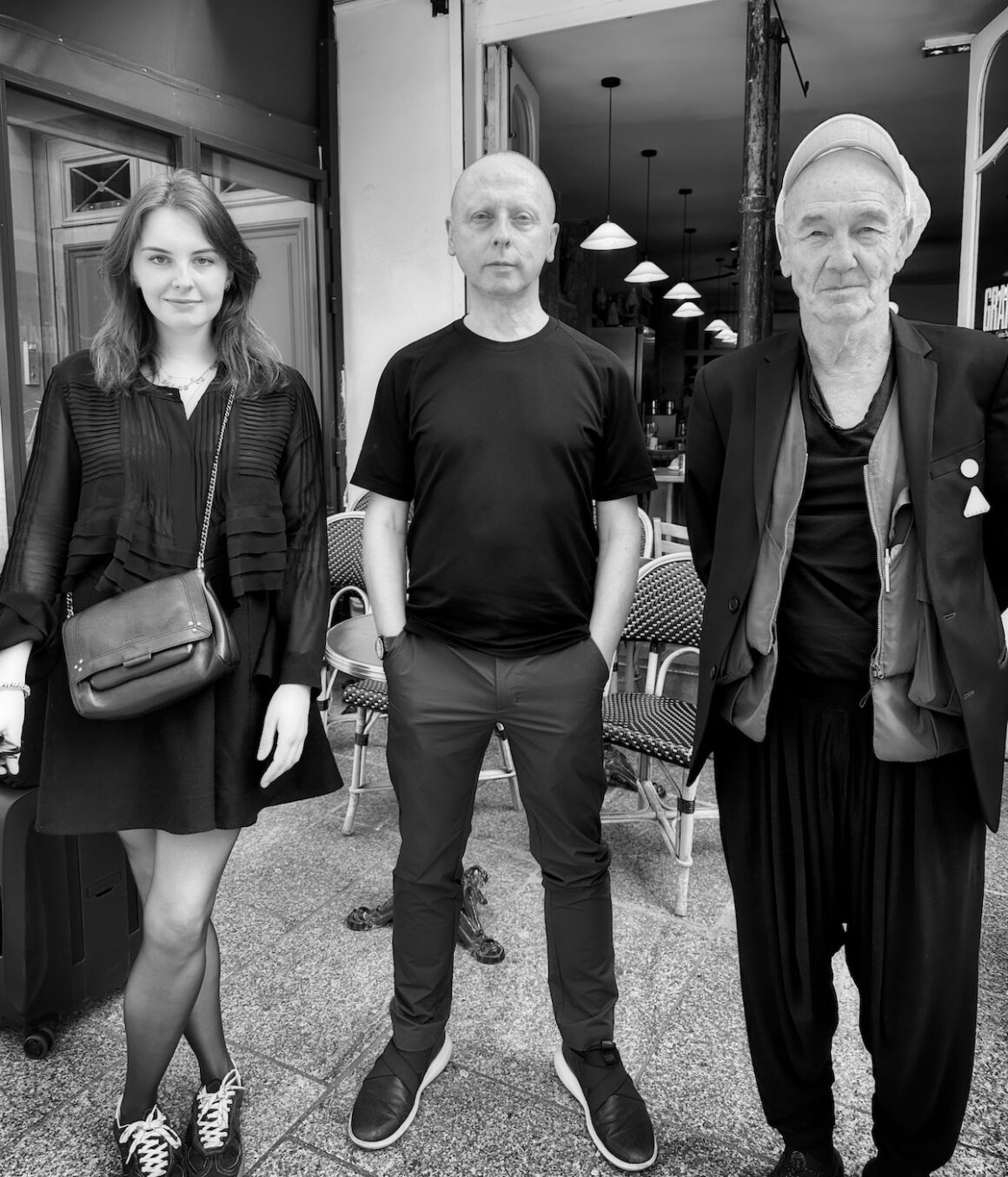 Black and white photo of three people standing in front of a cafe, with a young woman on the left wearing a black dress and sneakers, a middle-aged man in the middle wearing a black t-shirt and pants, and an older man on the right wearing a cap, blazer, and loose pants.
