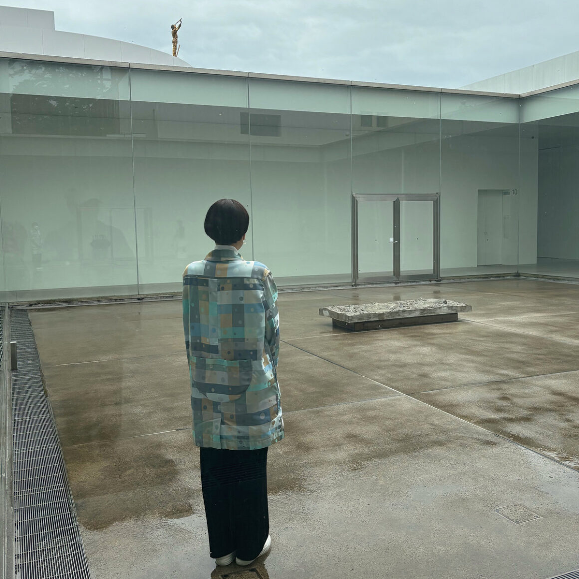 A female figure stands with arms at their sides, smartly dressed, with short black hair, in a square outside in Japan. She looks across the courtyeard at a bench of concrete, in a large empty quiet space