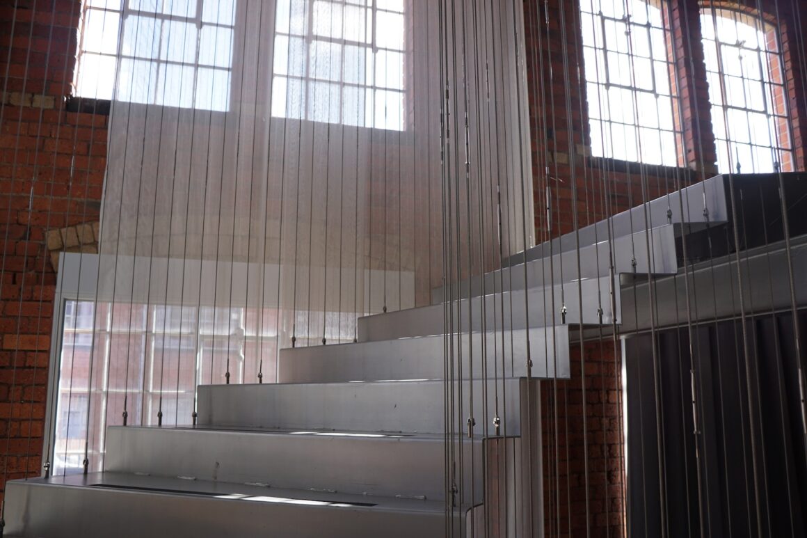 A stainless steel staircase, with windows behind it, in a large industrial space