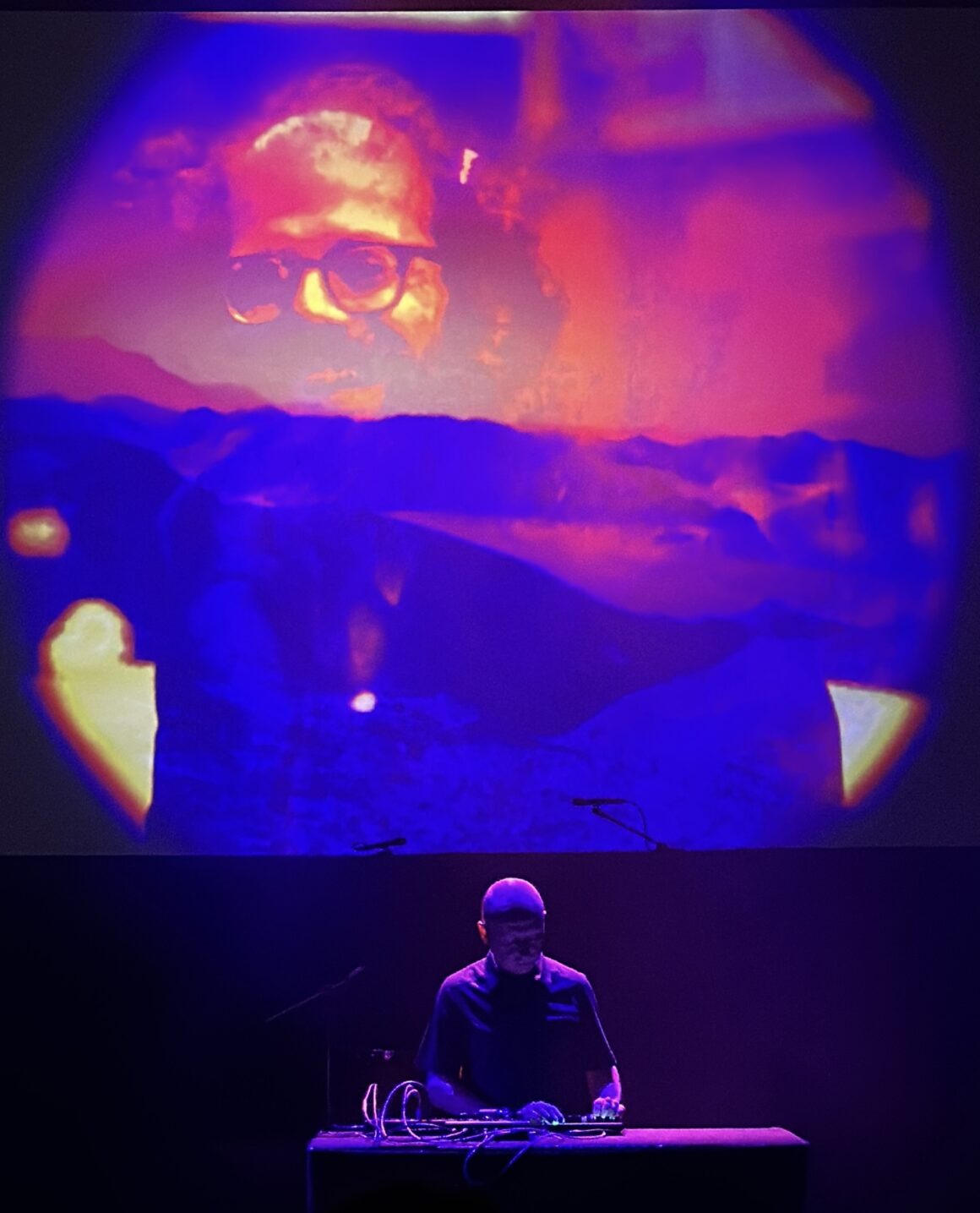 Musician performing live with an abstract colorful projection of a face and a landscape on a screen behind him in a dark venue.