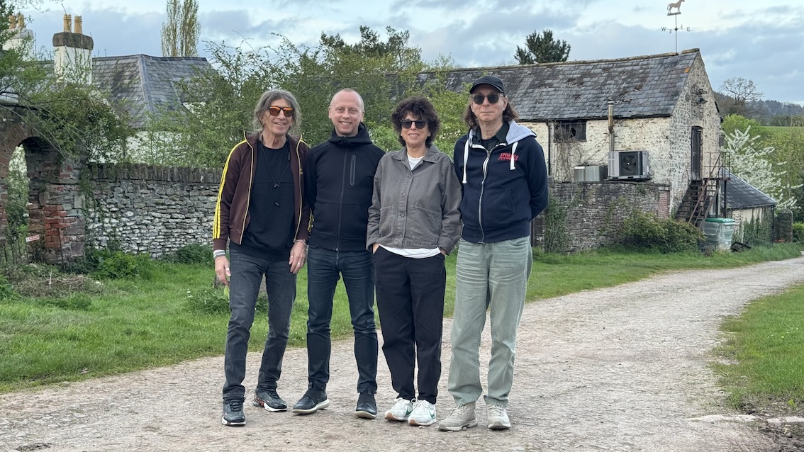 Four people stand in a row, three men and one woman, all smiling, standing on a country lane in Wales